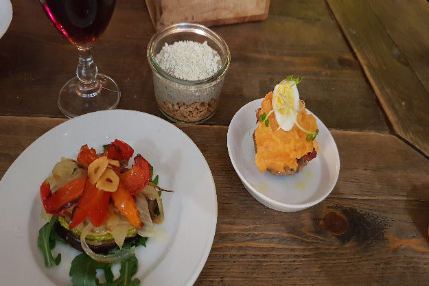 Drei verschiedene Tapas, darunter ein Nachtisch, angerichtet auf einem rustikalen Holztisch im spanischen Stil, begleitet von einem gefüllten Glas Rotwein – Szene aus dem Tapas-Kochkurs.