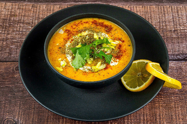 Leuchtend gelb-orange afghanische Dahlsuppe mit Joghurttopping, Petersilie und Staudensellerie in schwarzer Schale, serviert mit Zitronenscheiben – Afghanischer Kochkurs in Düsseldorf